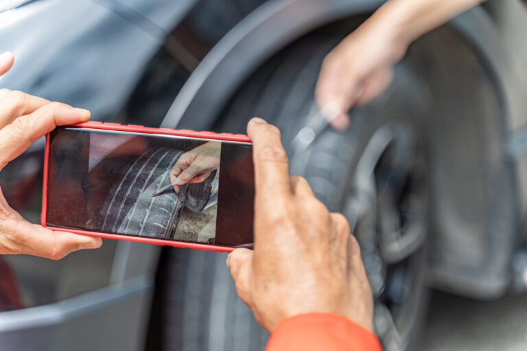 Close up hands of a car insurance agent taking a picture of a damage mark on the car tire while the custmer using his pen to point it out